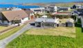 Aerial view of a single-storey cottage with a grey slate roof and large windows, beside a gravel driveway, with a sea inlet, green hills