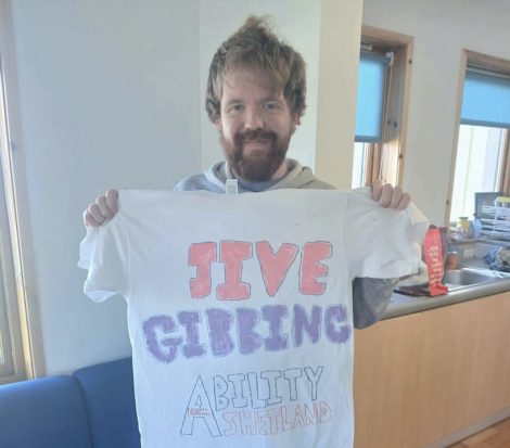 A man holding up a white T-shirt with the text “JIVE GIBBING ABILITY SHETLAND” written on the front.