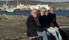 Three smiling men sit side by side on a stone harbour wall, with a white ferry and grey naval vessel moored in the harbour behind them