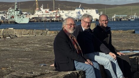 Three smiling men sit side by side on a stone harbour wall, with a white ferry and grey naval vessel moored in the harbour behind them