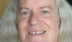 Close-up portrait of a smiling person with long, wavy silver-grey hair, showing a gap between their front teeth, against a light wooden