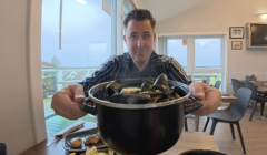 A smiling man in a striped navy jacket holds a large black pot of steamed mussels toward the camera in a bright café with large windows. A