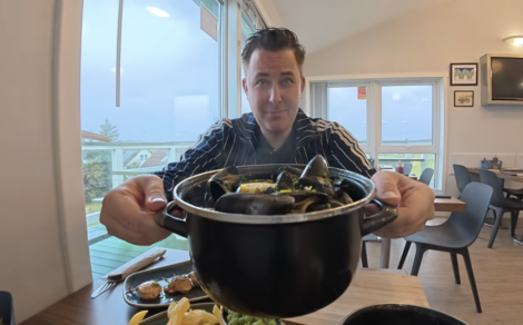 A smiling man in a striped navy jacket holds a large black pot of steamed mussels toward the camera in a bright café with large windows. A