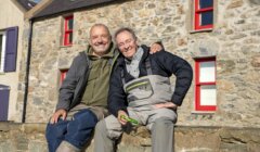 Two smiling men sit side by side on a stone wall in front of a stone house with red-framed windows. The man on the right wears grey waders