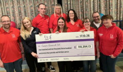Eight people in red Relay for Life shirts hold a giant cheque for £110,394.13 to Cancer Research UK in front of patterned curtains.