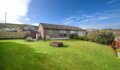 A single-storey bungalow with grey rendered walls and a dark tiled roof, set on a large, freshly mown lawn under a bright blue sky. A