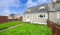 Grey rendered semi-detached house with a white front door, dormer windows and a dark slate roof, set behind a lawn and wooden fence under a