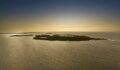 Aerial view of a low, green island surrounded by calm, golden-tinted sea under a sunset sky, with smaller islands visible on the horizon.
