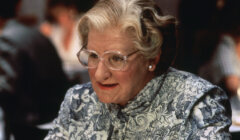 An older woman with curled silver-white hair, clear-framed glasses, pearl earrings and a black-and-white floral blouse sits at a table