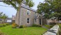 A two-storey stone house with a slate roof, white sash windows and a small front porch, set behind a lawn edged with flowering shrubs and a