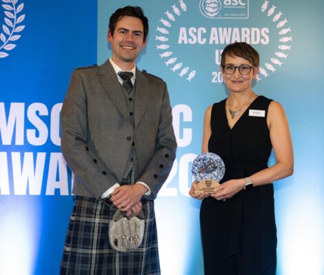 A smiling man in a grey Highland jacket and tartan kilt stands beside a woman in a black sleeveless outfit holding a blue-and-white award