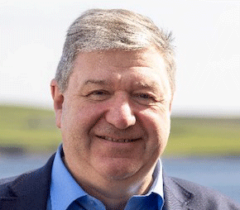 A man with short gray hair is smiling while wearing a blue shirt and a blazer. The background shows an out-of-focus natural landscape.