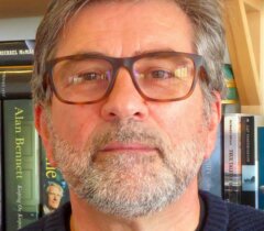 A bearded man with glasses is wearing a navy sweater. He stands in front of a bookshelf filled with books.