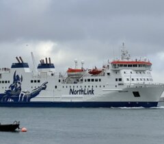 A large NorthLink ferry travels on the water under a cloudy sky, with a small boat anchored nearby in the foreground.