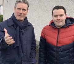 Two men standing outdoors in front of a light-colored wall; one is gesturing with his hand, both are wearing jackets suitable for cool weather.
