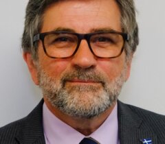 A middle-aged man with gray hair, glasses, and a beard, wearing a suit, light purple shirt, and navy tie with a small blue-and-white flag pin.